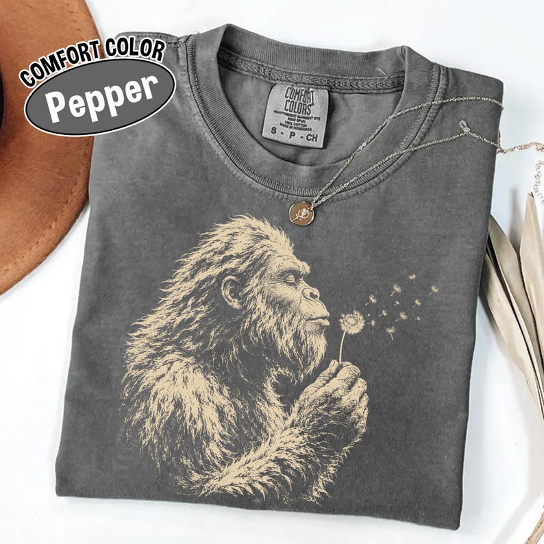 Puede incluir: Una camiseta Comfort Colors color pimienta con un gr&aacute;fico de una criatura peluda soplando un diente de le&oacute;n. La prenda tiene cuello redondo y una etiqueta que dice Comfort Colors S P CH. Un collar de cadena plateada con un dije redondo descansa encima.