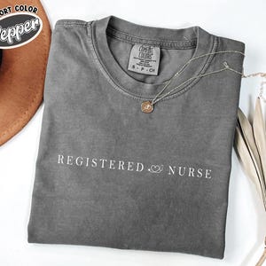 Peut inclure: Un t-shirt gris avec les mots "REGISTERED NURSE" imprimés en blanc. Un graphique de stéthoscope et de cœur se trouve entre les mots. Un collier en or avec un pendentif est sur le t-shirt. Le t-shirt est plié sur une surface blanche.