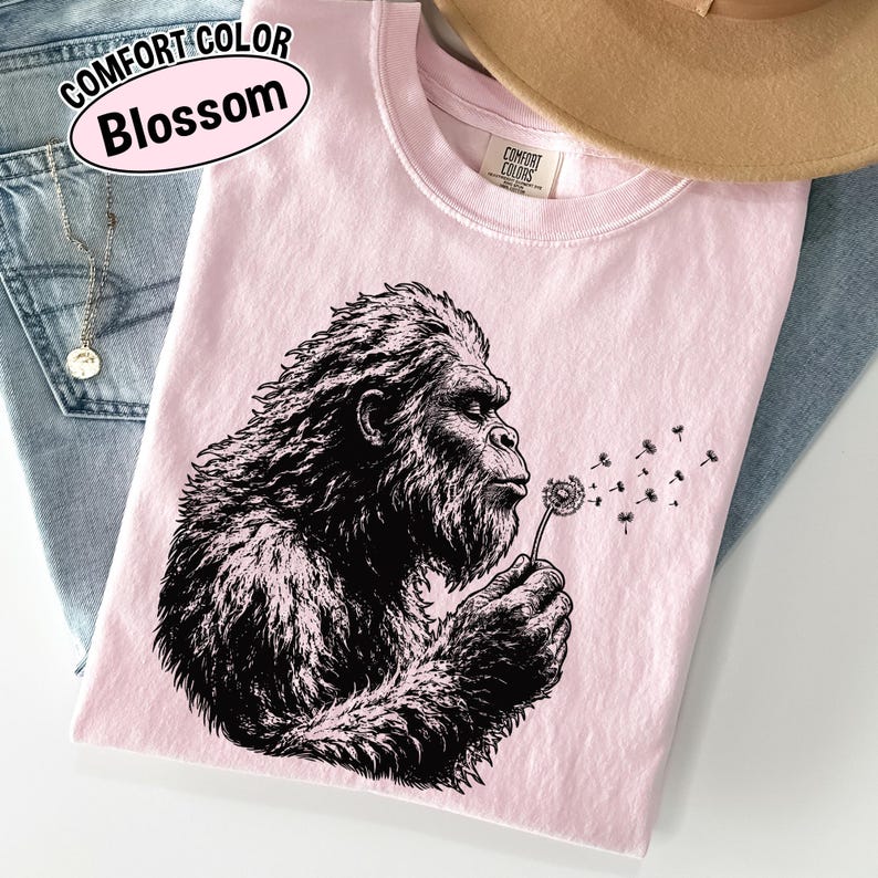 Puede incluir: Una camiseta rosa claro con un gr&aacute;fico negro de una criatura peluda soplando un diente de le&oacute;n, junto al texto COMFORT COLOR Blossom. La prenda tiene cuello redondo y una etiqueta interior con la inscripci&oacute;n COMFORT COLORS.