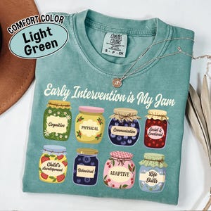 May include: Light green Comfort Colors t-shirt with the text "Early Intervention is My Jam." The shirt features illustrations of eight jam jars, each labeled with a different aspect of early intervention, such as "Cognitive" and "Life Skills."