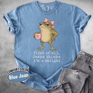 May include: A blue jean colored t-shirt featuring a graphic of a tan toad wearing a pink bow and holding a pink mug with a heart. Below the toad, the text reads: first of all, Jesus thinks i'm a delight. The shirt has a crew neck and short sleeves.