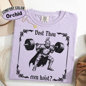 Może przedstawiać: Koszulka w kolorze orchidei z czarną grafiką rycerza podnoszącego sztangę, obramowaną ozdobami. Tekst "Dost Thou even hoist?" znajduje się nad i pod rycerzem. Koszulka ma metkę Comfort Colors.