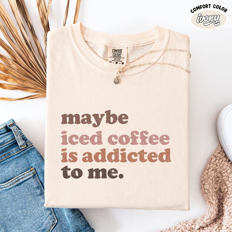 Peut inclure: T-shirt ivoire avec le texte "maybe iced coffee is addicted to me" en lettres marron et roses. Un collier argent&eacute; avec un pendentif repose sur le t-shirt. Le t-shirt est pli&eacute; sur une surface blanche &agrave; c&ocirc;t&eacute; d'un jean bleu.