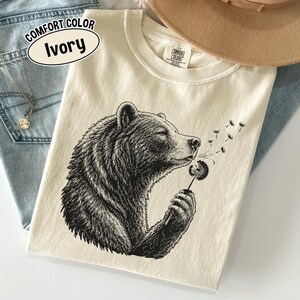 May include: An ivory t-shirt featuring a black ink-style illustration of a bear holding a dandelion. The bear is blowing on the dandelion, causing seeds to drift away. The shirt has a Comfort Colors label at the neck and a text bubble stating Comfort Color Ivory.