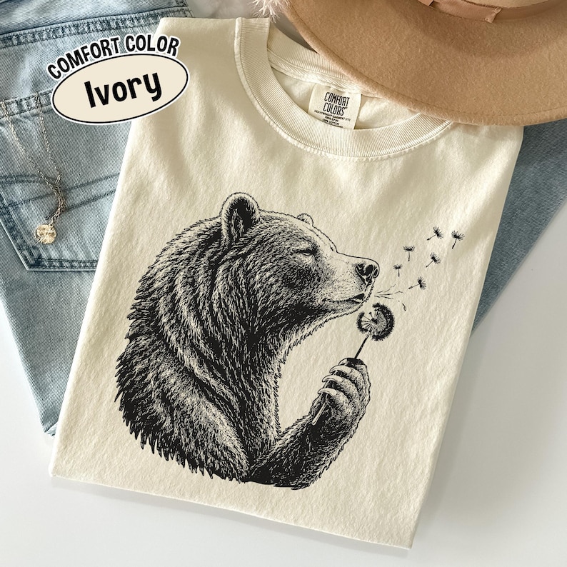 May include: An ivory t-shirt featuring a black ink-style illustration of a bear holding a dandelion. The bear is blowing on the dandelion, causing seeds to drift away. The shirt has a Comfort Colors label at the neck and a text bubble stating Comfort Color Ivory.