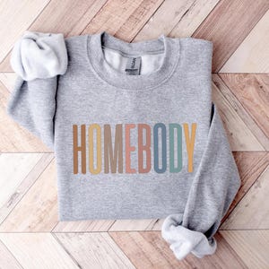 May include: A heather gray sweatshirt with the word "HOMEBODY" printed across the front in a colorful, retro font. The long sleeves are folded, and the sweatshirt is lying on a wooden surface.
