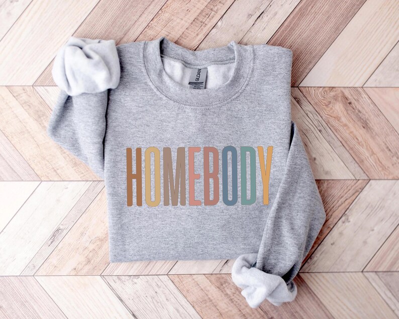 May include: A heather gray sweatshirt with the word "HOMEBODY" printed across the front in a colorful, retro font. The long sleeves are folded, and the sweatshirt is lying on a wooden surface.