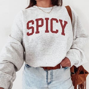 May include: A gray sweatshirt with the word "SPICY" printed in red on the front. The sweatshirt has a relaxed fit and long sleeves.