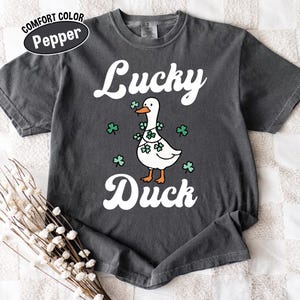 Puede incluir: Camiseta gris oscuro con las palabras "Lucky Duck" en escritura blanca. Un pato de dibujos animados con una guirnalda de tr&eacute;boles est&aacute; centrado debajo de la palabra "Lucky". La camiseta tiene una etiqueta Comfort Color Pepper.