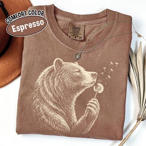 May include: An espresso-colored t-shirt featuring a white graphic print of a bear holding and blowing on a dandelion. The shirt has a crew neckline and a Comfort Colors brand label on the inside collar. The text on the label reads Comfort Colors.