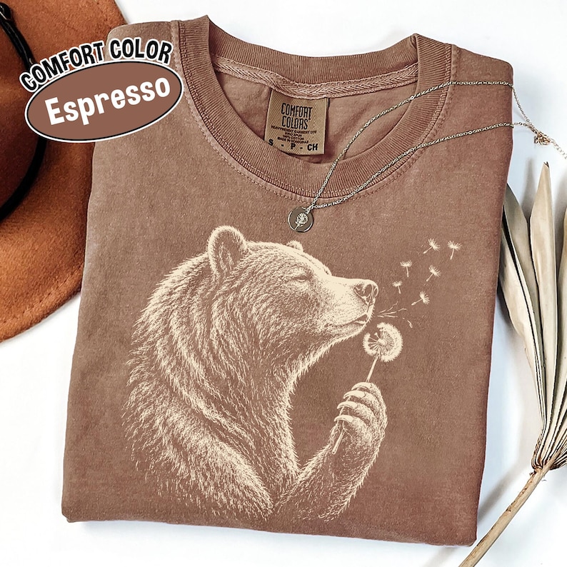 May include: An espresso-colored t-shirt featuring a white graphic print of a bear holding and blowing on a dandelion. The shirt has a crew neckline and a Comfort Colors brand label on the inside collar. The text on the label reads Comfort Colors.