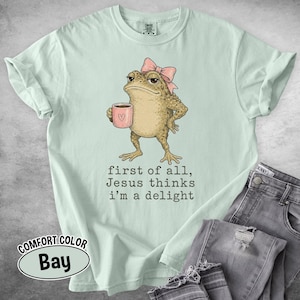 May include: A light mint green t-shirt featuring a graphic of a toad wearing a pink bow and holding a pink mug. The text below the toad reads, "first of all, Jesus thinks i'm a delight." The shirt has short sleeves and a crew neckline.