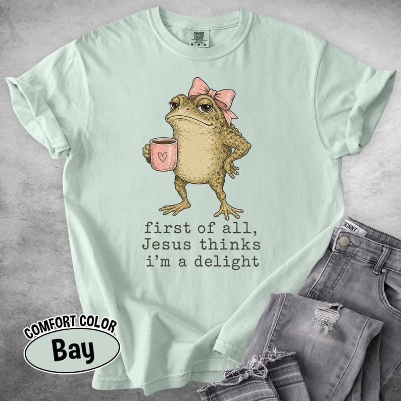 May include: A light mint green t-shirt featuring a graphic of a toad wearing a pink bow and holding a pink mug. The text below the toad reads, "first of all, Jesus thinks i'm a delight." The shirt has short sleeves and a crew neckline.