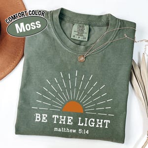 May include: A moss green t-shirt with the text "BE THE LIGHT" and "matthew 5:14" printed in white. The design includes a sunburst graphic with an orange sun. A gold necklace with a pendant is on the shirt.