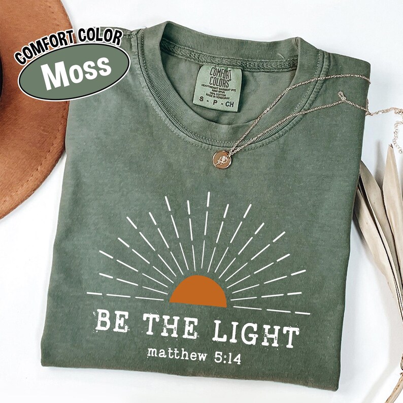 May include: A moss green t-shirt with the text "BE THE LIGHT" and "matthew 5:14" printed in white. The design includes a sunburst graphic with an orange sun. A gold necklace with a pendant is on the shirt.