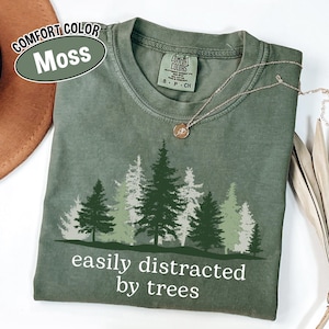 May include: A moss-colored t-shirt with a forest graphic and the text "easily distracted by trees." The design features various shades of green and white trees. A gold necklace with a pendant is draped on the shirt.