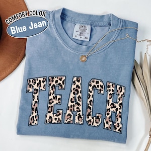 May include: A blue jean Comfort Colors t-shirt with the word "TEACH" in a leopard print design. The shirt has a crew neck and short sleeves. A gold necklace with a pendant is draped across the shirt.