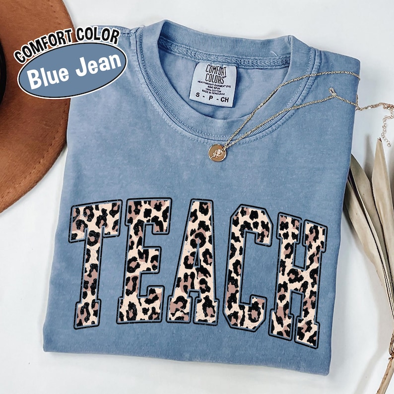 May include: A blue jean Comfort Colors t-shirt with the word "TEACH" in a leopard print design. The shirt has a crew neck and short sleeves. A gold necklace with a pendant is draped across the shirt.