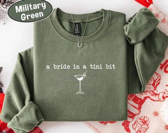 May include: Military green sweatshirt with the text "a bride in a tini bit" and a martini glass graphic. The long-sleeved crew neck shirt has a silver necklace and is displayed on a festive holiday background.