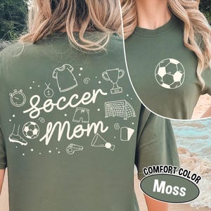K&ouml;nnte beinhalten: Ein moosgr&uuml;nes T-Shirt mit dem Schriftzug "soccer mom" in Schreibschrift und Fu&szlig;ball-Grafiken. Auf der Vorderseite befindet sich eine Fu&szlig;ballgrafik. Das Shirt ist mit "Comfort Color Moss" gekennzeichnet.