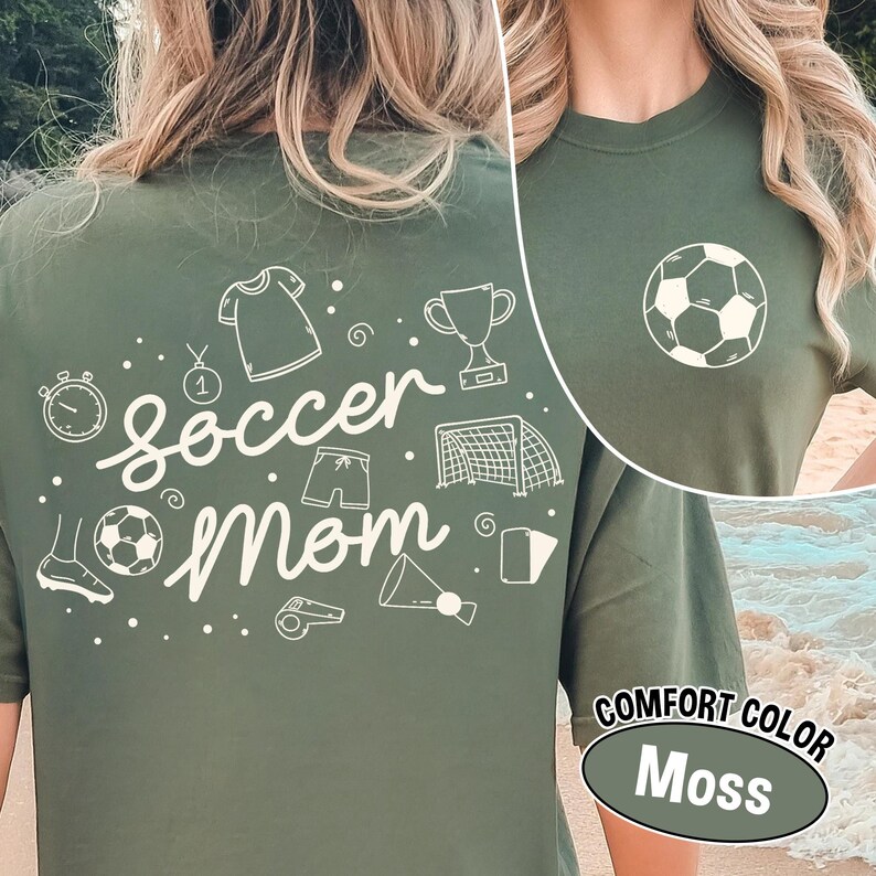 K&ouml;nnte beinhalten: Ein moosgr&uuml;nes T-Shirt mit dem Schriftzug "soccer mom" in Schreibschrift und Fu&szlig;ball-Grafiken. Auf der Vorderseite befindet sich eine Fu&szlig;ballgrafik. Das Shirt ist mit "Comfort Color Moss" gekennzeichnet.