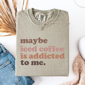 Peut inclure: Sweat-shirt vert sauge Comfort Colors avec le texte "maybe iced coffee is addicted to me" en lettres marron et roses. Un collier argent&eacute; avec un pendentif est pos&eacute; sur le v&ecirc;tement. Le sweat-shirt est pli&eacute; sur une surface blanche.