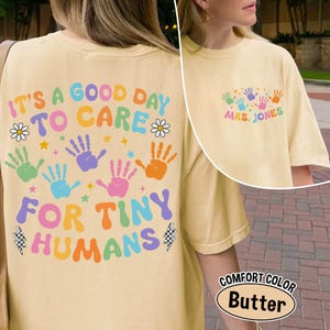 Puede incluir: Camiseta de color mantequilla con el texto "IT'S A GOOD DAY TO CARE FOR TINY HUMANS" en letras coloridas. El dise&ntilde;o incluye huellas de manos y flores. La camiseta tambi&eacute;n presenta el texto "MRS. JONES" en la parte delantera.