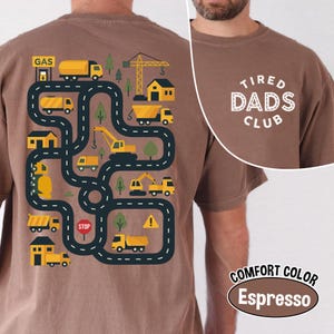 May include: Espresso-colored t-shirt with a playful road map design featuring yellow construction vehicles, buildings, and trees. The text "TIRED DADS CLUB" is printed on the upper right. The shirt is labeled "COMFORT COLOR".