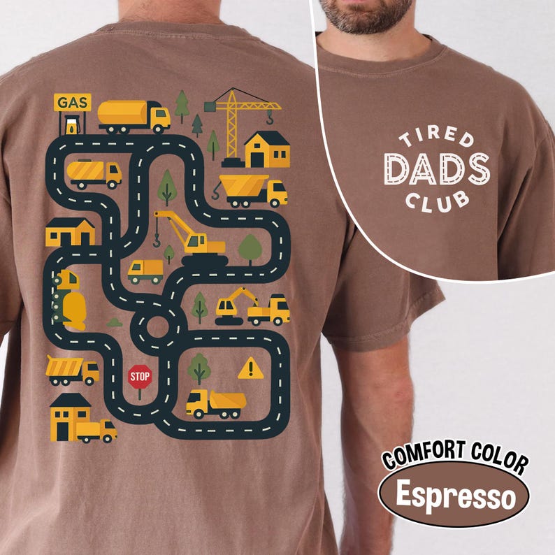 May include: Espresso-colored t-shirt with a playful road map design featuring yellow construction vehicles, buildings, and trees. The text "TIRED DADS CLUB" is printed on the upper right. The shirt is labeled "COMFORT COLOR".