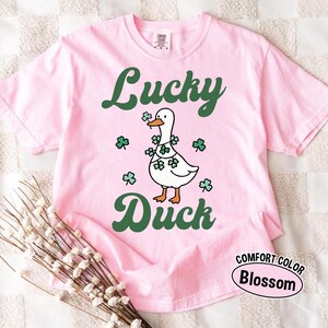 Puede incluir: Camiseta rosa claro con las palabras "Lucky Duck" en verde. Un pato blanco con una guirnalda de tr&eacute;boles es el motivo central. La camiseta est&aacute; etiquetada como "Comfort Color Blossom".