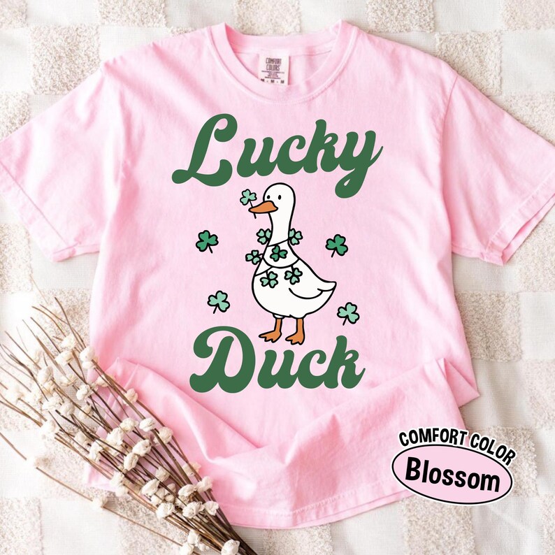 Puede incluir: Camiseta rosa claro con las palabras "Lucky Duck" en verde. Un pato blanco con una guirnalda de tr&eacute;boles es el motivo central. La camiseta est&aacute; etiquetada como "Comfort Color Blossom".