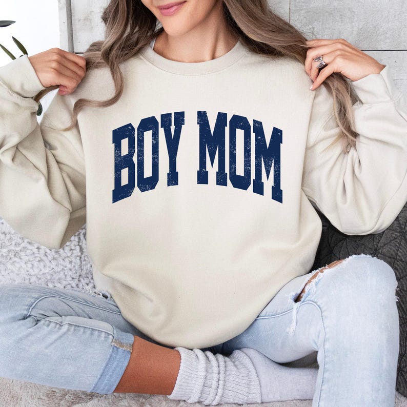 Puede incluir: Una sudadera color crema con el texto "BOY MOM" en azul marino, con letras de estilo universitario. La sudadera es de estilo casual, adecuada para el uso diario.
