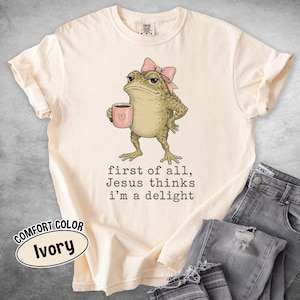 May include: An ivory short-sleeve t-shirt featuring a graphic of a green toad wearing a pink bow and holding a pink mug with a heart. Below the toad, the text reads: first of all, Jesus thinks i'm a delight. The shirt has a crew neckline.