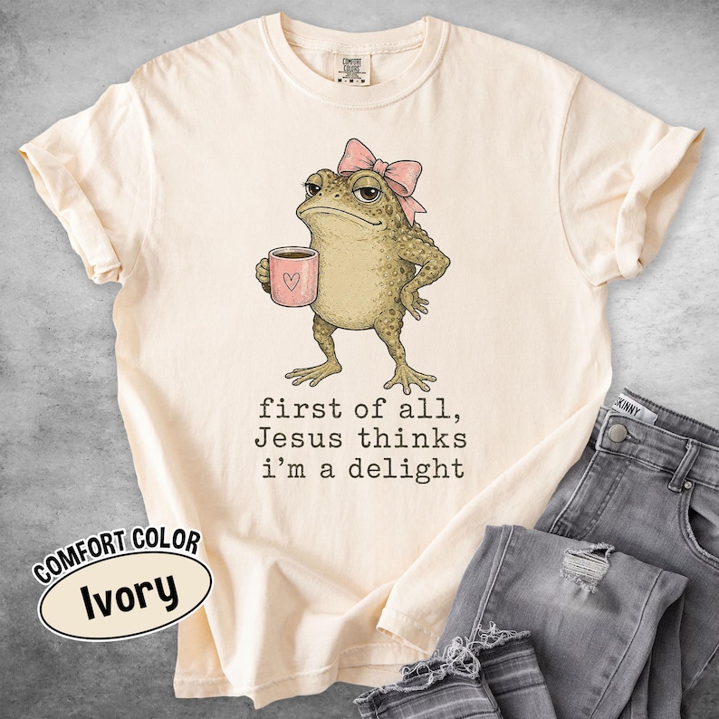 May include: An ivory short-sleeve t-shirt featuring a graphic of a green toad wearing a pink bow and holding a pink mug with a heart. Below the toad, the text reads: first of all, Jesus thinks i'm a delight. The shirt has a crew neckline.