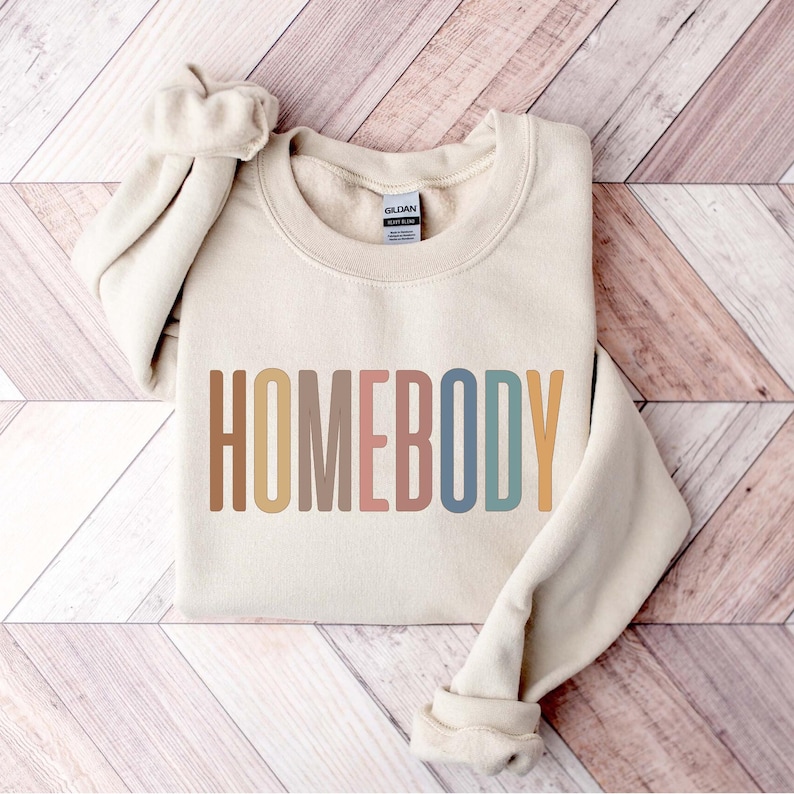 May include: A beige sweatshirt with the word "HOMEBODY" printed across the front in a rainbow of colors. The long sleeves are cuffed at the wrists. The sweatshirt is folded on a wooden surface.