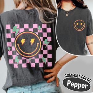 May include: Dark gray t-shirt featuring a smiley face with lightning bolts on the front and back. The back design includes a pink and black checkered pattern. The shirt has the words "COMFORT COLOR Pepper" printed on it.
