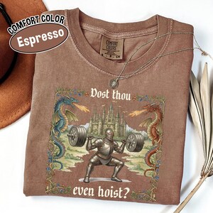 May include: Espresso-colored Comfort Colors t-shirt featuring a knight lifting weights with a castle backdrop and dragons. The shirt has the text "Dost thou even hoist?" and a Comfort Colors tag.