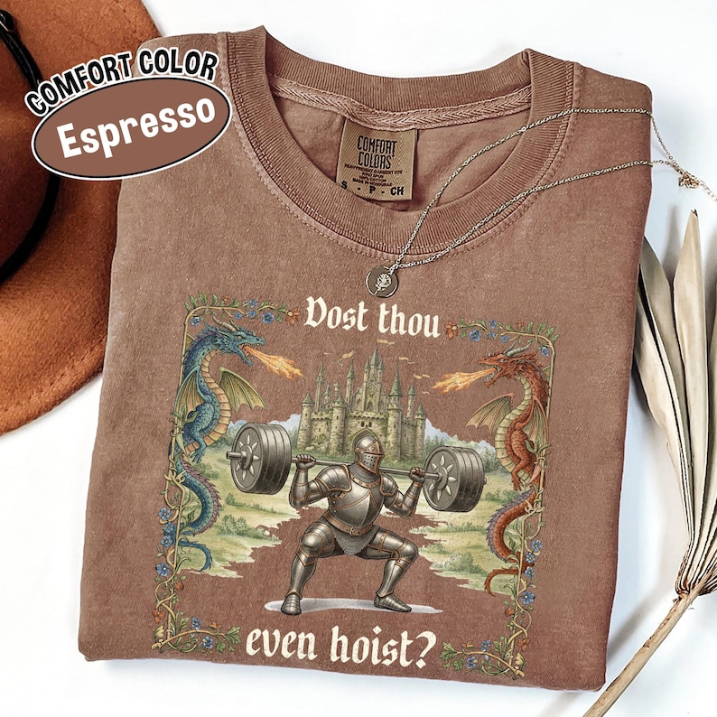 May include: Espresso-colored Comfort Colors t-shirt featuring a knight lifting weights with a castle backdrop and dragons. The shirt has the text "Dost thou even hoist?" and a Comfort Colors tag.
