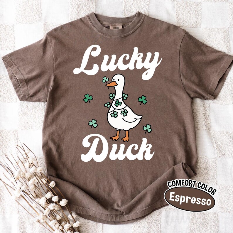 Puede incluir: Camiseta color espresso con las palabras "Lucky Duck" en escritura blanca. Un pato de dibujos animados con una guirnalda de tr&eacute;boles aparece en el dise&ntilde;o. La camiseta es de la marca Comfort Color.