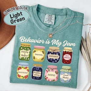 May include: Light green Comfort Colors t-shirt with the phrase "Behavior is My Jam" and illustrations of jam jars labeled with positive attributes like "Empathy & Respect" and "Growth Mindset". A silver necklace is draped across the shirt.