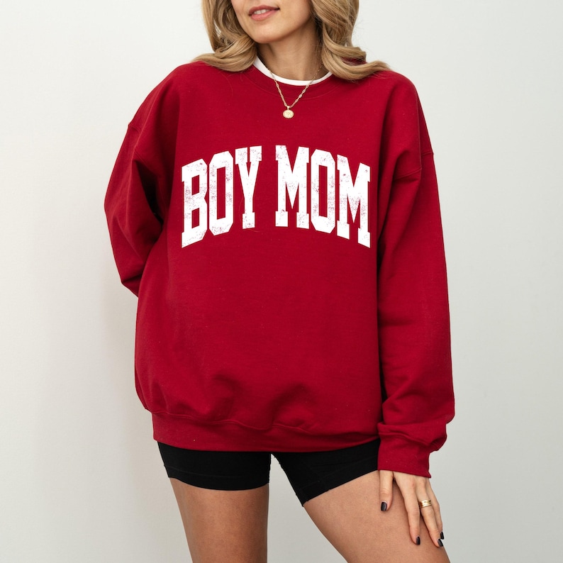 Puede incluir: Una sudadera roja con el texto "BOY MOM" en letras blancas grandes. La sudadera tiene cuello redondo y mangas largas. La modelo lleva un pantal&oacute;n corto negro.
