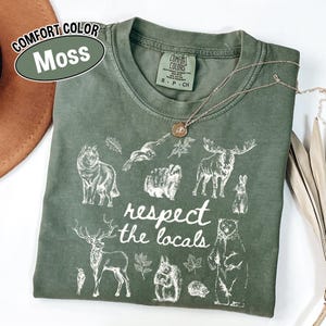 May include: A moss green Comfort Colors t-shirt with a white graphic of woodland animals and the text "respect the locals." The shirt has a crew neck and short sleeves. A gold necklace is draped across the shirt.