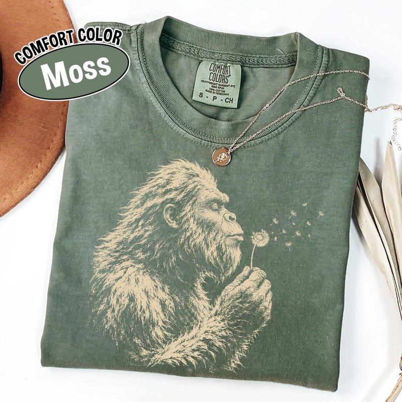Puede incluir: Una camiseta verde musgo con un estampado gr&aacute;fico de una criatura peluda soplando un diente de le&oacute;n. La prenda tiene cuello redondo y una etiqueta de la marca Comfort Colors en el interior. Una fina cadena dorada descansa sobre la tela.