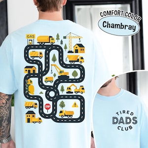 May include: Light blue t-shirt with a colorful graphic of a road with construction vehicles, trees, and buildings. The text "COMFORT COLOR Chambray" is printed on the upper right. The lower right has the text "TIRED DADS CLUB".