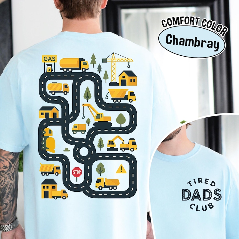 May include: Light blue t-shirt with a colorful graphic of a road with construction vehicles, trees, and buildings. The text "COMFORT COLOR Chambray" is printed on the upper right. The lower right has the text "TIRED DADS CLUB".