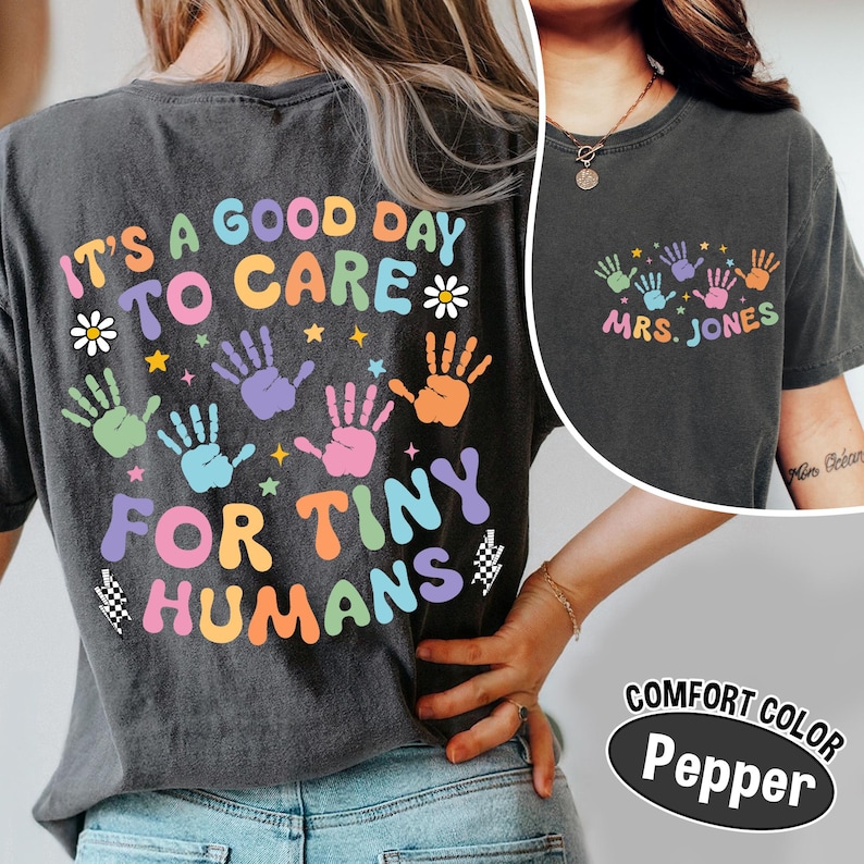 Puede incluir: Camiseta gris oscuro Comfort Colors con el texto "IT'S A GOOD DAY TO CARE FOR TINY HUMANS" en letras coloridas y huellas de manos. La camiseta tambi&eacute;n presenta el nombre "MRS. JONES" en la parte delantera. La camiseta es de color pimienta.