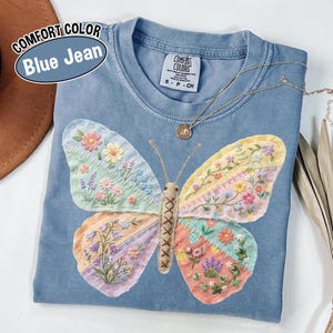 May include: Blue jean Comfort Colors t-shirt with a large, embroidered butterfly design. The butterfly wings feature a patchwork of pastel colors and floral patterns. A gold necklace with a pendant rests on the shirt.