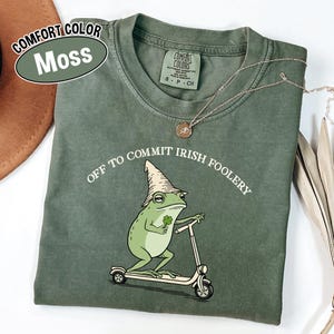 May include: A moss green Comfort Colors t-shirt with the phrase "Off to Commit Irish Foolery" and a cartoon frog riding a scooter. The frog wears a hat and holds a four-leaf clover. A silver necklace is draped across the shirt.