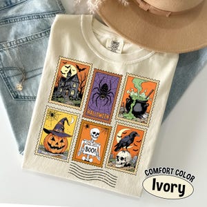 May include: Ivory-colored Comfort Colors t-shirt featuring a collection of Halloween-themed postage stamp designs. The stamps depict a haunted house, spider, cauldron, jack-o'-lantern, skeleton, and raven. The shirt includes the text "COMFORT COLOR" and "Ivory".
