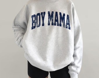 May include: A light gray crewneck sweatshirt featuring the text BOY MAMA in a dark navy blue, distressed, collegiate-style font arched across the chest. The long-sleeved pullover has a relaxed fit and is paired with black shorts.